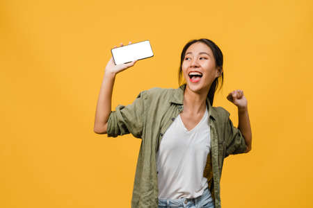 Young Asia lady show empty smartphone screen with positive expression, smiles broadly, dressed in casual clothing feeling happiness on yellow background. Mobile phone with white screen in female hand.の写真素材