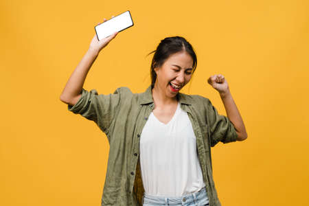 Young Asia lady show empty smartphone screen with positive expression, smiles broadly, dressed in casual clothing feeling happiness on yellow background. Mobile phone with white screen in female hand.の写真素材