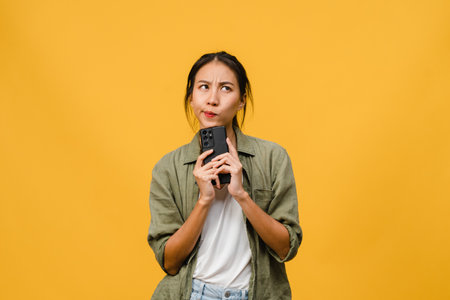 Thinking dreaming young Asia lady using phone with positive expression, dressed in casual cloth feeling happiness and stand isolated on yellow background. Happy adorable glad woman rejoices success.の写真素材