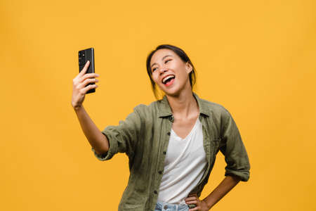Smiling adorable Asian female making selfie photo on smart phone with positive expression in casual clothing and stand isolated on yellow background. Happy adorable glad woman rejoices success.の写真素材