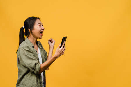 Surprised young Asia lady using mobile phone with positive expression, smiles broadly, dressed in casual clothing and stand isolated on yellow background. Happy adorable glad woman rejoices success.の写真素材