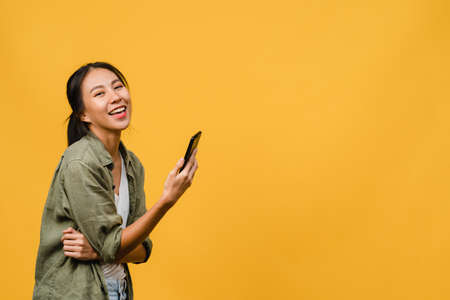 Surprised young Asia lady using mobile phone with positive expression, smile broadly, dressed in casual clothing and looking at camera on yellow background. Happy adorable glad woman rejoices success.の写真素材