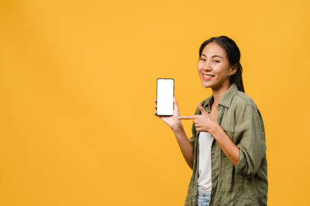 Young Asia lady show empty smartphone screen with positive expression, smiles broadly, dressed in casual clothing feeling happiness on yellow background. Mobile phone with white screen in female hand.の写真素材