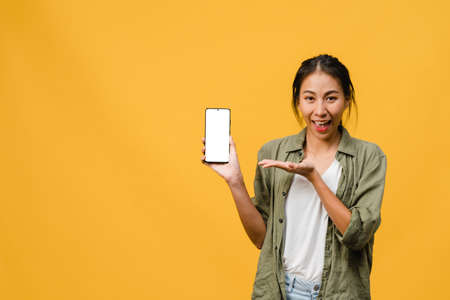 Young Asia lady show empty smartphone screen with positive expression, smiles broadly, dressed in casual clothing feeling happiness on yellow background. Mobile phone with white screen in female hand.の写真素材