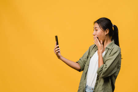 Young Asia lady using phone with positive expression, smiles broadly, dressed in casual clothing feeling happiness and stand isolated on yellow background. Happy adorable glad woman rejoices success.の写真素材