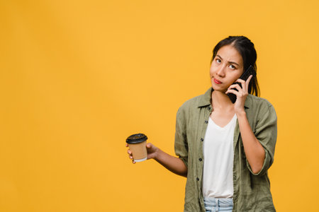Young Asia lady talk by phone and hold coffee cup with negative expression, excited screaming, cry emotional angry in casual cloth and stand isolated on yellow background. Facial expression concept.の写真素材