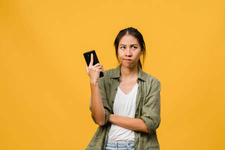 Thinking dreaming young Asia lady using phone with positive expression, dressed in casual cloth feeling happiness and stand isolated on yellow background. Happy adorable glad woman rejoices success.の写真素材