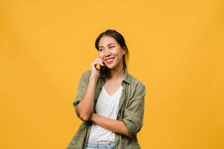Young Asia lady talk by phone with positive expression, smile broadly, dressed in casual clothing feeling happiness and stand isolated on yellow background. Happy adorable glad woman rejoices success.の写真素材