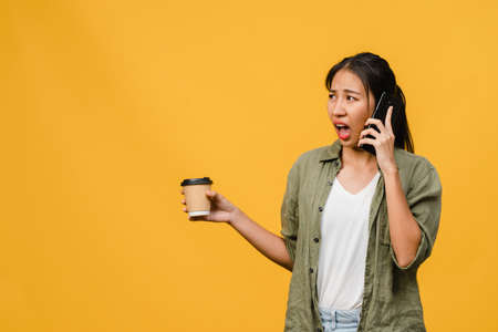 Young Asia lady talk by phone and hold coffee cup with negative expression, excited screaming, cry emotional angry in casual cloth and stand isolated on yellow background. Facial expression concept.の写真素材