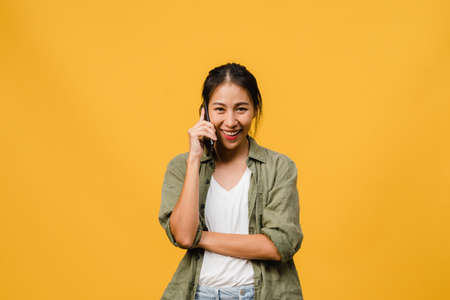 Young Asia lady talk by phone with positive expression, smile broadly, dressed in casual clothing feeling happiness and stand isolated on yellow background. Happy adorable glad woman rejoices success.の写真素材