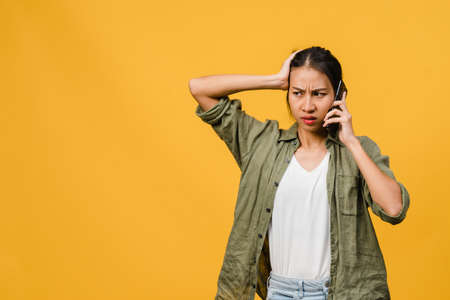 Young Asia lady talk by phone with negative expression, excited screaming, cry emotional angry in casual cloth and stand isolated on yellow background with blank copy space. Facial expression concept.の写真素材