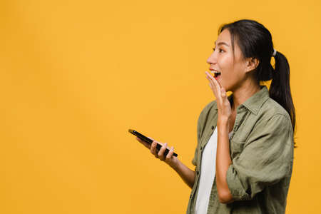 Young Asia lady using phone with positive expression, smiles broadly, dressed in casual clothing feeling happiness and stand isolated on yellow background. Happy adorable glad woman rejoices success.の写真素材