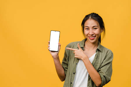 Young Asia lady show empty smartphone screen with positive expression, smiles broadly, dressed in casual clothing feeling happiness on yellow background. Mobile phone with white screen in female hand.の写真素材