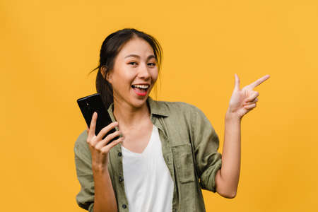 Young Asian lady using mobile phone with cheerful expression, shows something amazing at blank space in casual cloth and looking at camera isolated over yellow background. Facial expression concept.の写真素材