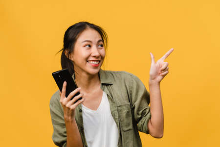 Portrait of young Asian lady using mobile phone with cheerful expression, show something amazing at blank space in casual clothing and stand isolated over yellow background. Facial expression concept.の写真素材