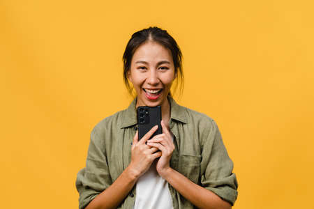 Surprised young Asia lady using mobile phone with positive expression, smile broadly, dressed in casual clothing and looking at camera on yellow background. Happy adorable glad woman rejoices success.の写真素材
