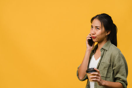 Young Asia lady talk by phone and hold coffee cup with negative expression, excited screaming, cry emotional angry in casual cloth and stand isolated on yellow background. Facial expression concept.の写真素材