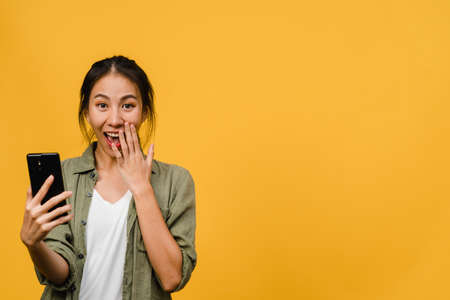 Surprised young Asia lady using mobile phone with positive expression, smiles broadly, dressed in casual clothing and stand isolated on yellow background. Happy adorable glad woman rejoices success.の写真素材