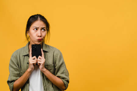 Thinking dreaming young Asia lady using phone with positive expression, dressed in casual cloth feeling happiness and stand isolated on yellow background. Happy adorable glad woman rejoices success.の写真素材
