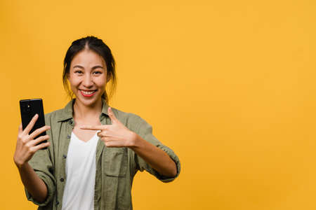 Young Asian lady using mobile phone with cheerful expression, shows something amazing at blank space in casual cloth and looking at camera isolated over yellow background. Facial expression concept.の写真素材