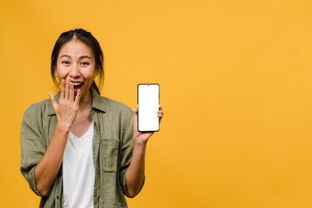 Young Asia lady show empty smartphone screen with positive expression, smiles broadly, dressed in casual clothing feeling happiness on yellow background. Mobile phone with white screen in female hand.の写真素材