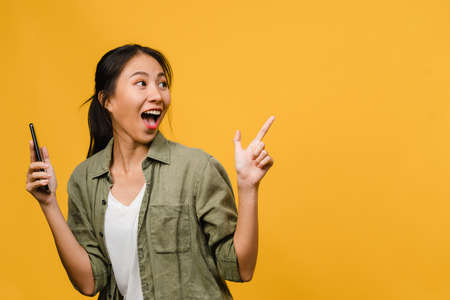 Portrait of young Asian lady using mobile phone with cheerful expression, show something amazing at blank space in casual clothing and stand isolated over yellow background. Facial expression concept.の写真素材