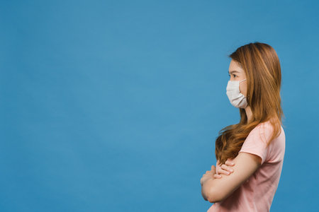 Young Asia girl wearing medical face mask with dressed in casual cloth and looking at blank space isolated on blue background. Self-isolation, social distancing, quarantine for corona virus preventionの写真素材