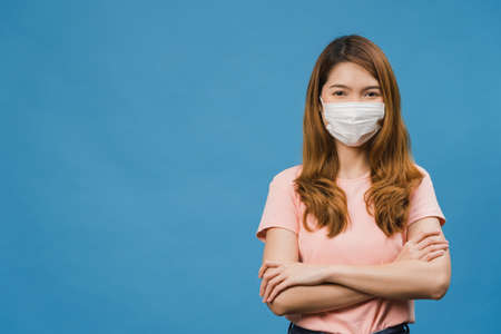 Young Asia girl wearing medical face mask with arms crossed, dressed in casual cloth and looking at camera isolated on blue background. Self-isolation, social distancing, quarantine for corona virus.の写真素材