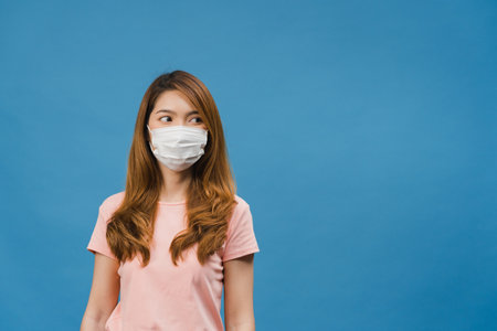 Young Asia girl wearing medical face mask with dressed in casual cloth and looking at blank space isolated on blue background. Self-isolation, social distancing, quarantine for corona virus preventionの写真素材