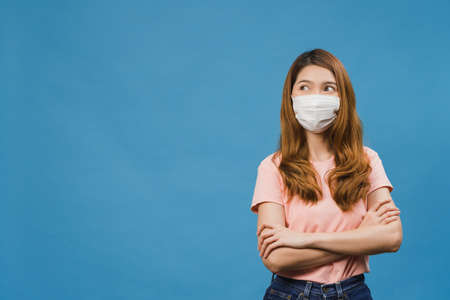 Young Asia girl wearing medical face mask with dressed in casual cloth and looking at blank space isolated on blue background. Self-isolation, social distancing, quarantine for corona virus preventionの写真素材