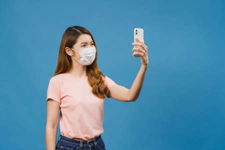 Smiling adorable Asian female wearing medical face mask making selfie photo on smart phone with positive expression in casual clothing and stand isolated on blue background.の写真素材