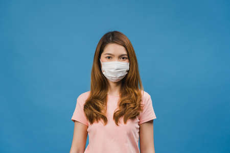 Young Asia girl wearing medical face mask with dressed in casual clothing and looking at camera isolated on blue background. Self-isolation, social distancing, quarantine for corona virus prevention.の写真素材