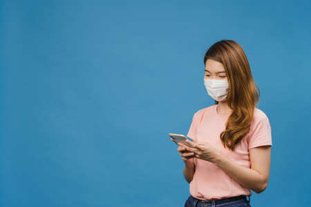 Young Asia girl wearing medical face mask using mobile phone with dressed in casual clothing isolated on blue background. Self-isolation, social distancing, quarantine for corona virus prevention.の写真素材