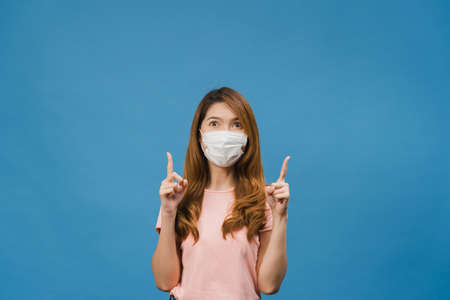 Young Asia girl wear medical face mask shows something at blank space with dressed in casual cloth and look at camera isolated on blue background. Social distancing, quarantine for corona virus.の写真素材