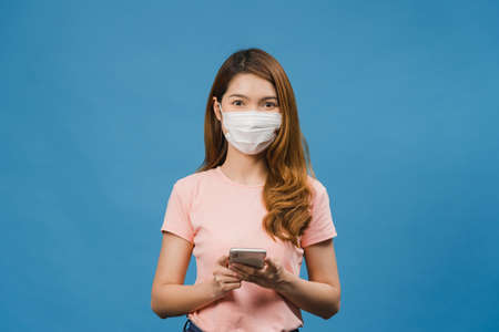 Young Asia girl wearing medical face mask using mobile phone with dressed in casual clothing isolated on blue background. Self-isolation, social distancing, quarantine for corona virus prevention.の写真素材