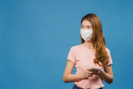 Young Asia girl wearing medical face mask using mobile phone with dressed in casual clothing isolated on blue background. Self-isolation, social distancing, quarantine for corona virus prevention.の写真素材