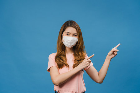 Young Asia girl wearing medical face mask shows something at blank space with dressed in casual cloth and looking at camera isolated on blue background. Social distancing, quarantine for corona virus.の写真素材