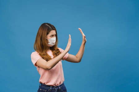 Young Asia lady wear medical face mask doing stop sing with palm of hand with negative expression and looking at camera isolated on blue background. Social distancing, quarantine for corona virus.の写真素材