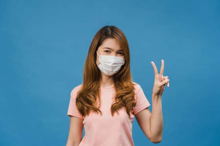Young Asia girl wearing medical face mask showing peace sign, encourage with dressed in casual cloth and looking at camera isolated on blue background. Social distancing, quarantine for corona virus.の写真素材