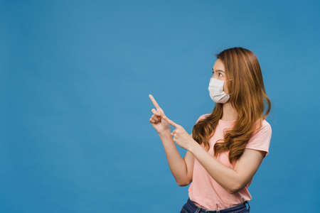 Young Asia girl wear medical face mask shows something at blank space with dressed in casual cloth and look at camera isolated on blue background. Social distancing, quarantine for corona virus.の写真素材