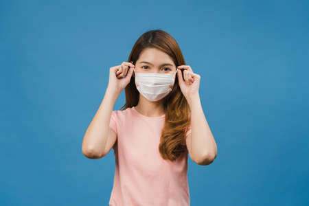 Young Asia girl wearing medical face mask with dressed in casual clothing and looking at camera isolated on blue background. Self-isolation, social distancing, quarantine for corona virus prevention.の写真素材