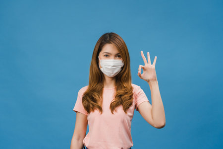 Young Asia girl wearing medical face mask gesturing ok sign with dressed in casual cloth and look at camera isolated on blue background. Self-isolation, social distancing, quarantine for corona virus.の写真素材
