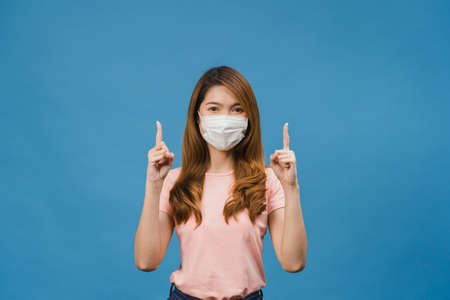 Young Asia girl wearing medical face mask shows something at blank space with dressed in casual cloth and looking at camera isolated on blue background. Social distancing, quarantine for corona virus.の写真素材