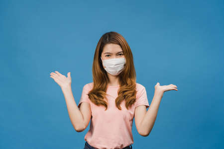 Young Asia girl wearing medical face mask showing peace sign, encourage with dressed in casual cloth and looking at camera isolated on blue background. Social distancing, quarantine for corona virus.の写真素材