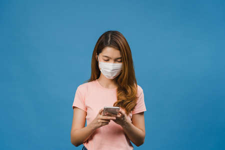 Young Asia girl wearing medical face mask using mobile phone with dressed in casual clothing isolated on blue background. Self-isolation, social distancing, quarantine for corona virus prevention.の写真素材