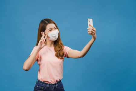 Smiling adorable Asian female wearing medical face mask making selfie photo on smart phone with positive expression in casual clothing and stand isolated on blue background.の写真素材
