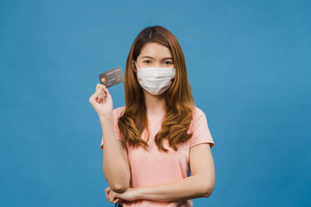 Young Asia lady wearing medical face mask show credit bank card with positive expression, smiles broadly, dressed in casual clothing feeling happiness and stand isolated on blue background.の写真素材