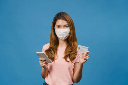 Young Asia lady wearing medical face mask using phone and credit bank card with positive expression, smiles broadly, dressed in casual clothing and stand isolated on blue background.の写真素材