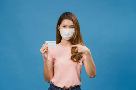 Young Asia lady wearing medical face mask show credit bank card with positive expression, smiles broadly, dressed in casual clothing feeling happiness and stand isolated on blue background.の写真素材