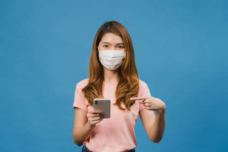 Young Asia girl wearing medical face mask using mobile phone with dressed in casual clothing isolated on blue background. Self-isolation, social distancing, quarantine for corona virus prevention.の写真素材